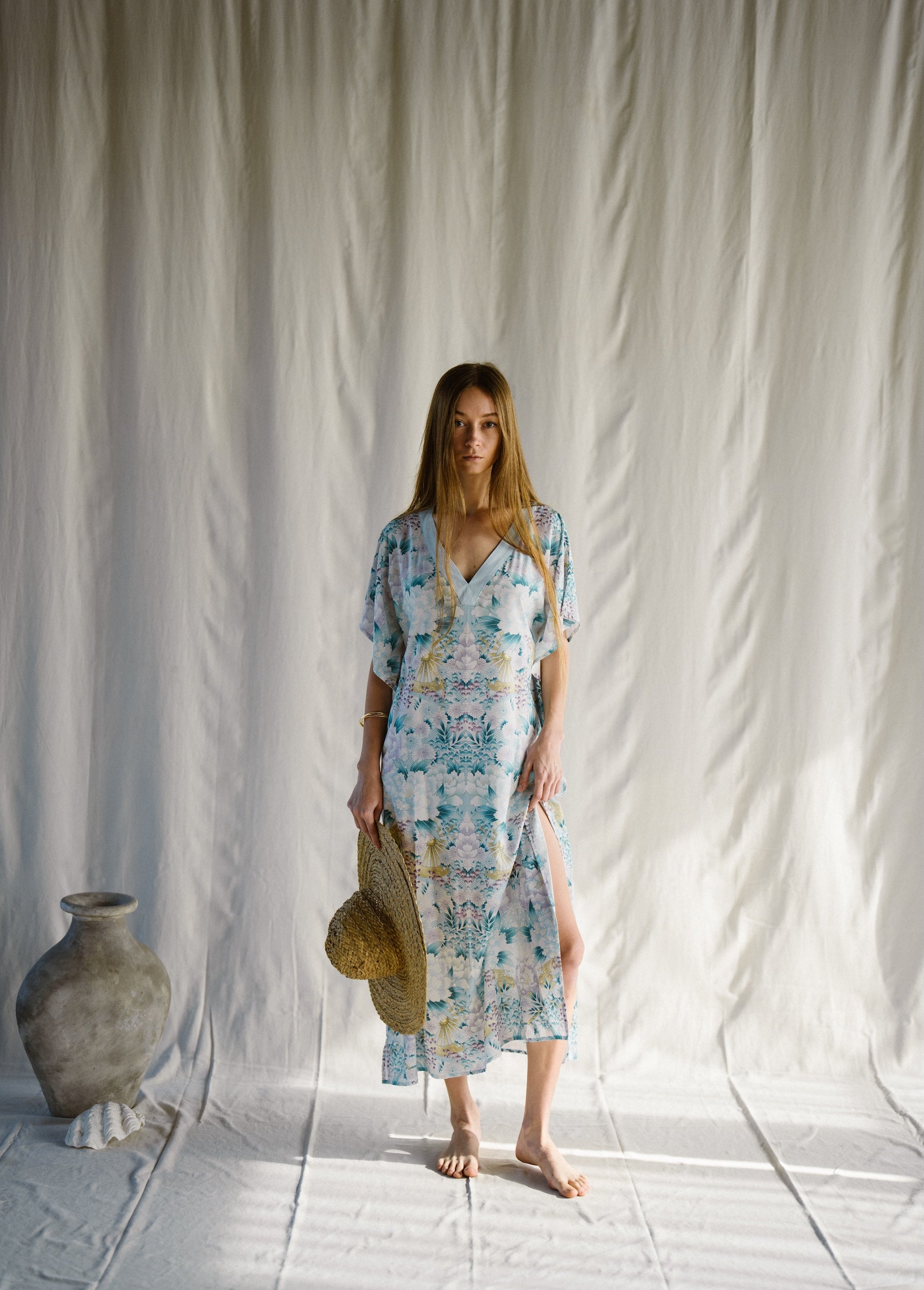 Woman in a floral long kaftan dress standing against a white curtain with a textured floor.