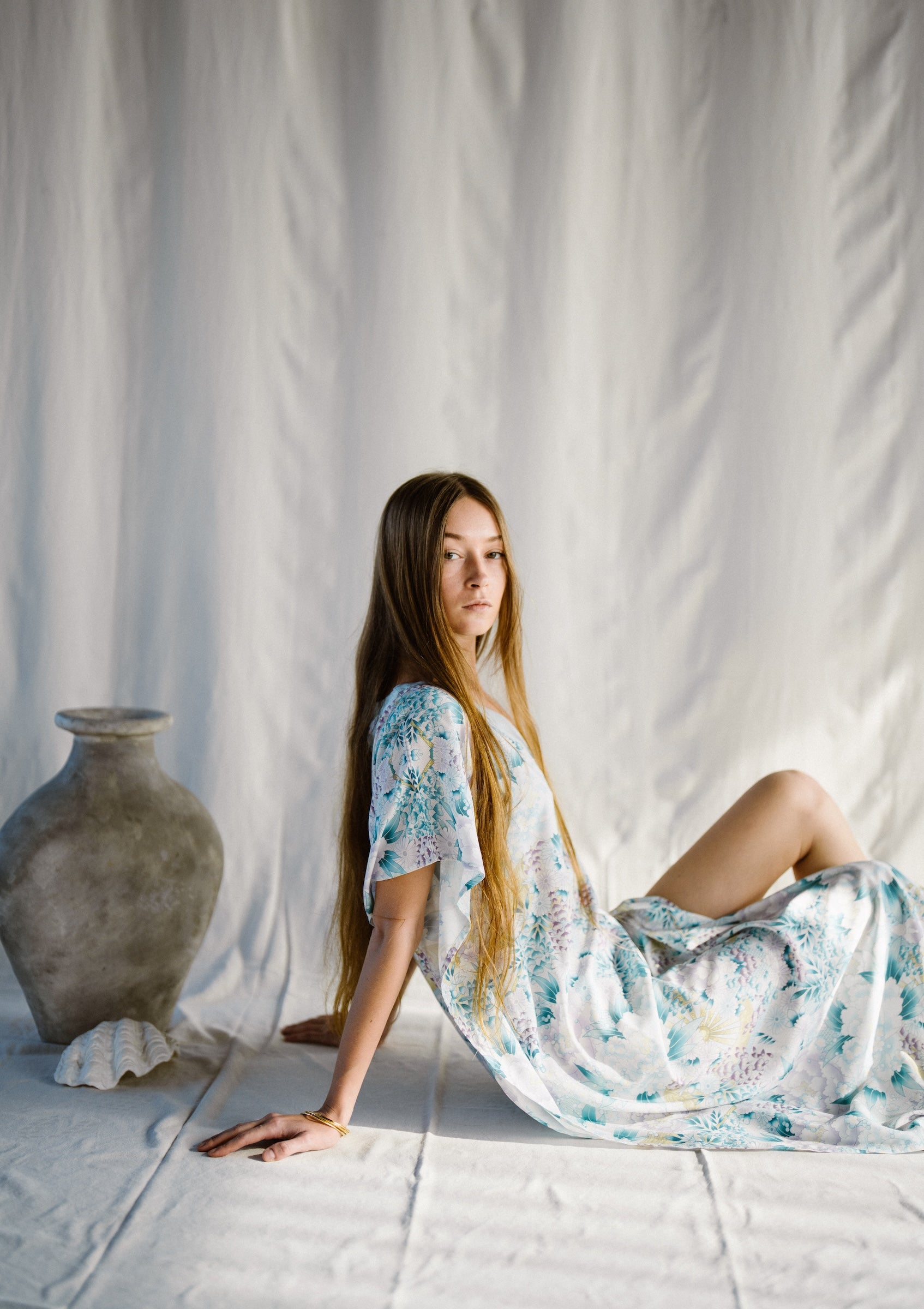 Woman in a floral kaftan dress sitting on the floor against a white curtain with a vase beside her.